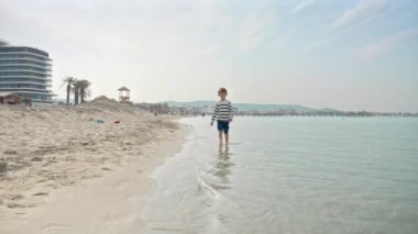 Mutlu gülümseyen yalın ayaklı çocuk, sakin dalgalar içinde kumların üzerinde yürüyor. Tatil, tatil, doğada hafta sonu.