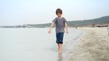 Çıplak ayaklı çocuk kumsalda koşar, su sıçratır, çocukluğun kaygısız ruhu, seyahat, mutluluk ve macera arayışı.