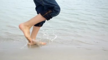 Kumlu sahilde koşan ve deniz suyu sıçratan çocuğun ayakları yavaş hareket ediyor. Seyahat, tatil, tatil, gezi, yolculuk ve yaz tatili kavramı
