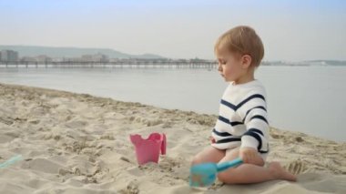 Gülümseyen çocuk kumsalda eğleniyor ve kum kazmak için kürek kullanıyor. Aile yolculuğunun ve hafta sonu kaçamağının mutluluğu ve heyecanı.