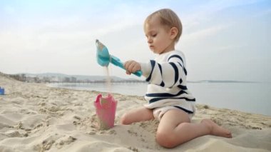 Mutlu erkek bebek yaz tatili boyunca kumlu sahilde kürekle oynuyor. Aile seyahatlerinin ve tatillerin kaygısız neşesi
