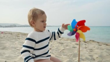 Kumlu deniz sahilinde oturan ve renkli fırıldak tekeriyle oynayan sevimli erkek bebek portresi. Yaz tatili veya hafta sonu çocuk mutluluğu kavramı