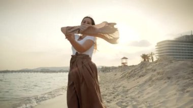 Genç bir kadının yavaş hareketi kumsalda renkli ipek bir atkıyı döndürüyor ve gülümsüyor ve rüzgara gülüyor. Mutluluğa, seyahate ve boş zamanlara vurgu