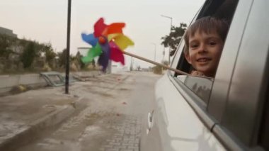 Açık araba camından dışarı bakan ve elinde dönen renkli fırıldak tutan gülümseyen çocuğun portresi..