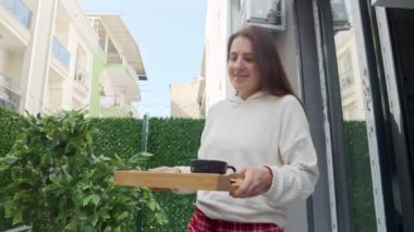 Kahvaltı tepsisiyle terasa doğru yürüyen mutlu, gülümseyen bir kadın, masanın yanında oturuyor ve dizüstü bilgisayarla çalışıyor. Serbest meslek anlayışı, evden çalışmak, uzak bir iş, dışarıda yemek yemek.