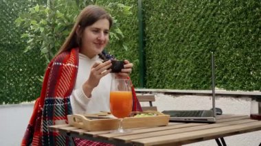 Ekoseyle kaplı genç bir kadın açık terasta dizüstü bilgisayarda çalışıyor ve kahve içiyor. Serbest meslek anlayışı, evden çalışmak, uzak bir iş, dışarıda yemek yemek.