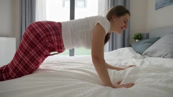 一个穿着睡衣的年轻女人倒在一个大窗户旁边的床上 脸上挂着欢乐的笑容 周末休闲和舒适家庭的概念 — 图库视频影像