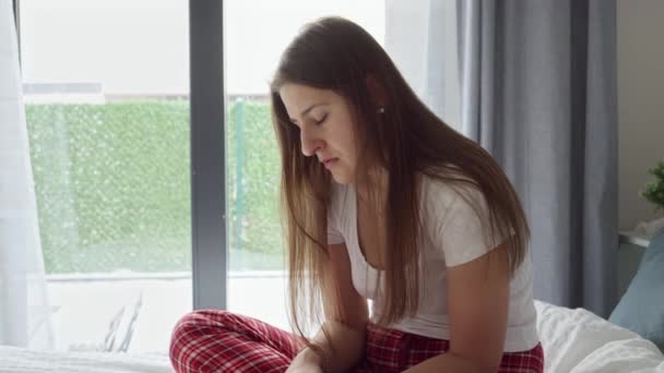 大きな窓の横にあるベッドの上に座っているパジャマ姿の動揺と孤独な若い女性 — ストック動画