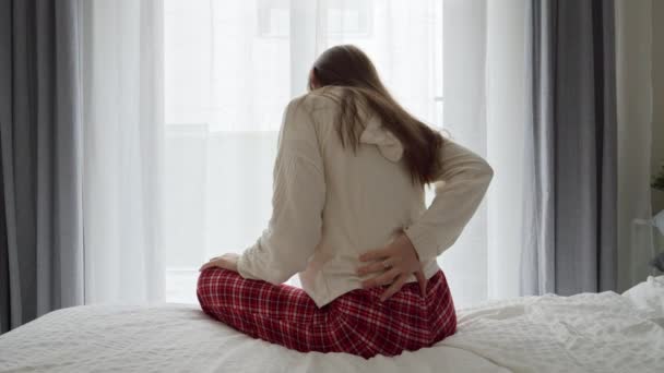 パジャマを着た若い女性がベッドサイドに座り 背中の痛みに苦しんでいます 痛みに関する問題の概念 — ストック動画
