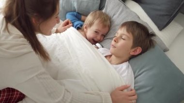 İki oğlunu battaniyeyle sarıp okşayan şefkatli genç bir anne. Aile mutluluğu kavramı, evde dinlenmek, yatakta eğlenmek, ebeveynler ve neşeli çocuklar..