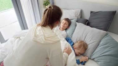 Genç ve sevgi dolu anne, sabahları yatakta uyuyan iki oğlunu okşuyor. Aile mutluluğu kavramı, evde dinlenmek, yatakta eğlenmek, ebeveynler ve neşeli çocuklar..
