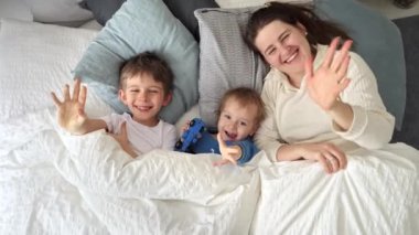 Genç mutlu anne iki oğluyla yatakta uzanıyor ve kameraya el sallıyor. Aile mutluluğu kavramı, evde dinlenmek, yatakta eğlenmek, ebeveynler ve neşeli çocuklar.