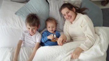 Oğulları battaniyenin altında yatıp kameraya gülümsemekten kurtulan mutlu bir anne. Aile mutluluğu kavramı, evde dinlenmek, yatakta eğlenmek, ebeveynler ve neşeli çocuklar..