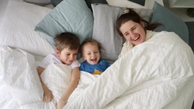 İki çocuk battaniyenin altında saklanıp onu atarken mutlu ve gülen bir aile. Aile mutluluğu kavramı, evde dinlenmek, yatakta eğlenmek, ebeveynler ve neşeli çocuklar..