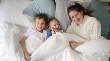 Yatakta iki oğlu kameraya el sallayan neşeli genç bir anne. Bu, aile mutluluğunun, evde kaliteli zaman geçirmenin ve ebeveynler ve çocuklar arasındaki neşeli anların özünü yakalar.