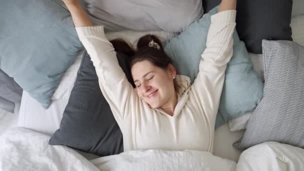 リラックスした若い女性は柔らかい枕の上に横になり 午前中にベッドでストレッチ 快適さ リラクゼーション 健康的な睡眠と新しい一日の良いスタートの概念 — ストック動画