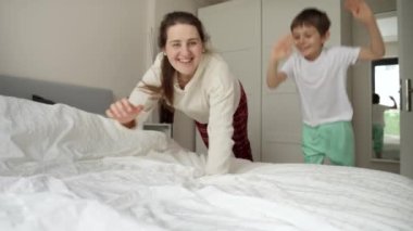 Annesi pijamayla koşan ve yatak odasındaki yumuşak yatağa düşen neşeli bir çocuk. Aile mutluluğu kavramı, evde dinlenmek, yatakta eğlenmek, ebeveynler ve neşeli çocuklar..