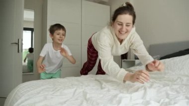 Yavaşça gülümseyen, pijamalı oğluyla birlikte yatağa atlayan ve sarılan bir anne. Aile mutluluğu kavramı, evde dinlenmek, yatakta eğlenmek, ebeveynler ve neşeli çocuklar..
