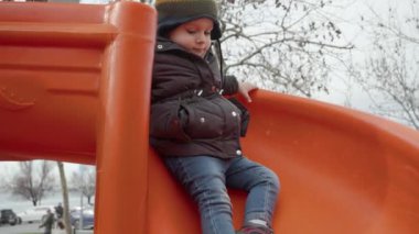 Çocuk parkında kaydırağından kayarak inen, sıcak şapkalı ve ceketli, gülümseyen bir çocuğun yavaş hareketi..