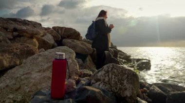 Dolly, genç bayan yürüyüşçüyü deniz kıyısındaki kayalıklarda dikilirken ve sıcak çay içerken okyanusun üzerinde gün batımını izlerken çekiyor.