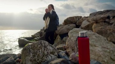 Gün batımında kayalık deniz kıyısında dinlenen ve soğuk rüzgarlı bir günde sıcak çay içen genç bir kadın. Kışın turizm, seyahat, seyahat, tatil ve tatil kavramı
