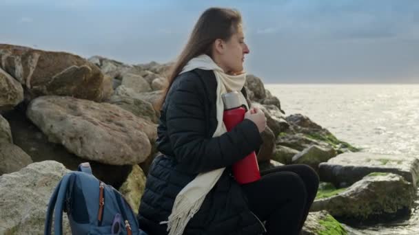 長い髪をした若い女性が海岸の大きな岩の上に座って 海の上に嵐の天気を見ています 冬の観光 休暇の概念 — ストック動画