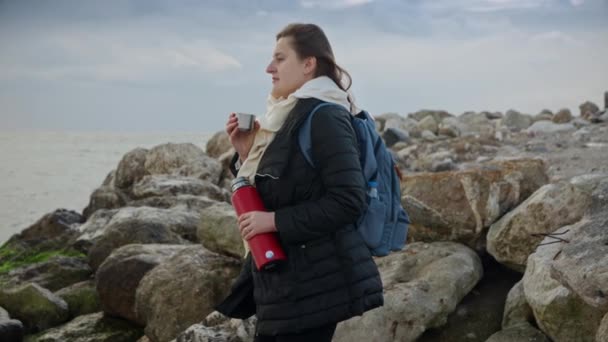 若い女性は海でリラックスし 熱いお茶の魔法瓶を持ち 野生で頑丈な海岸線を賞賛します またはライフスタイルコンテンツでの使用に最適です — ストック動画