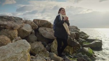 Elinde çay fincanıyla gülümseyen genç bir kadın soğuk deniz suyunun üzerinde gün batımını seyrediyor. Kışın turizm, seyahat, seyahat, tatil ve tatil kavramı