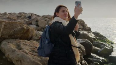 Genç bir kadın, kayalık sahilde dururken akıllı telefonuyla fotoğraf çekerek kış denizinin güzelliğini yakalıyor. Seyahat ve tatil görüntüleri için mükemmel.