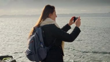 Kayalık sahilde sırt çantalı genç bir kadın akıllı telefonuyla soğuk kış denizinin fotoğraflarını çekiyor. Turizm ve seyahat videoları için mükemmel.