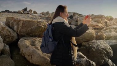 Genç bir kadın kayalıklarda dururken akıllı telefonuyla fotoğraf çekerek kış denizinin güzelliğini yakalıyor. Tatil ve seyahat için ideal