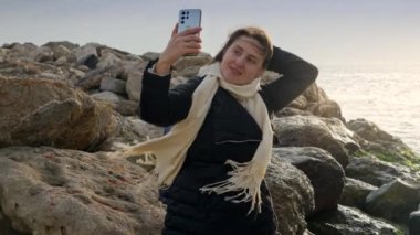 Genç bir kadın kış denizinde kayalık sahilde dururken akıllı telefonunu kullanarak fotoğraf çekiyor. Seyahat ve tatil görüntüleri için mükemmel.