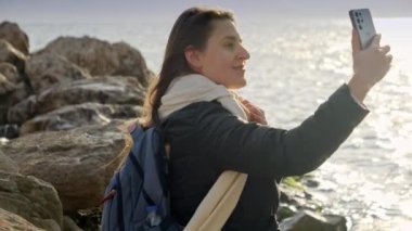 Gülümseyen kadın Susnet 'teki fırtınalı kış denizinin fotoğraflarını çekiyor kayaların üzerinde dururken akıllı telefonunu kullanıyor. Seyahat ve turizm için mükemmel..