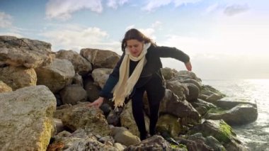 Sırt çantalı genç bir kadın soğuk bir günde gün batımında deniz kıyısında kaygan kayalıklarda tırmanıyor ve yürüyor. Kışın turizm, seyahat, seyahat, tatil ve tatil kavramı.