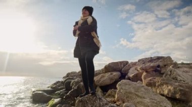 Uçurumda duran genç bir kadın, sırtında bir sırt çantasıyla denizin üzerinde kış günbatımını seyrediyor. Macera, keşif ve seyahat kavramı.