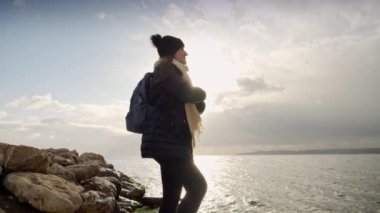 Şapkalı ve atkılı genç bir kadın uçurumun kenarında sırt çantasıyla, denizin üzerinde gün batımını seyrediyor. Seyahat, macera ve yeni hedefler keşfetme kavramı.