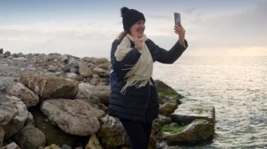 Deniz kıyısındaki uçurumlarda duran şapkalı ve atkılı gülümseyen kadın akıllı telefon kullanarak görüntülü konuşma yapıyor. Kışın turizm, seyahat, seyahat, tatil ve tatil kavramı.