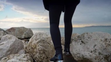 Sırt çantalı genç bir kadın gün batımında deniz kıyısındaki kayalıklara tırmanıyor. Kış turizmi, seyahat ve macera konsepti