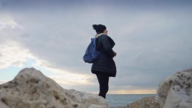 Sırt çantalı genç bir kadın kayalık bir uçurumun kenarından gün batımına bakıyor. Bir macera, keşif ve kış manzaralarının güzelliğinin tadını çıkarma kavramı.