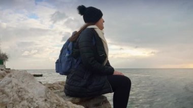 Sırt çantalı genç bir bayan yürüyüşçü uçurumda oturuyor ve rüzgarlı bir günde soğuk deniz dalgalarının üzerinde gün batımını seyrediyor. Macera kavramı, yürüyüş, seyahat, yolculuk ve doğa keşfi.