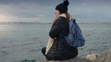 Sırt çantalı bir kadın maceracı, kayalık bir uçurumda mola veriyor, soğuk ve rüzgarlı denizin üzerinde güzel bir gün batımını seyrediyor. Macera, seyahat, yolculuk ve kendini doğaya daldırma kavramı.