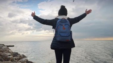 Sırt çantalı genç bir kadın, deniz kıyısındaki uçurumun kenarında uzanmış elleriyle soğuk okyanus dalgalarının tadını çıkarıyor. Yürüyüş, macera, doğa, keşif ve turizm kavramı.