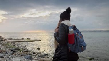 Sırt çantası ve termosuyla kadın yürüyüşçü, soğuk ve rüzgarlı bir havada kayalık bir deniz sahilinde yürüyor. Seyahat, turizm ve macera projeleri için mükemmel.