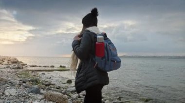 Sırt çantası ve termosuyla soğuk ve rüzgarlı bir günde engebeli kıyı şeridini keşfeden genç bir kadın. Macera, keşif ve açık hava seyahati kavramı