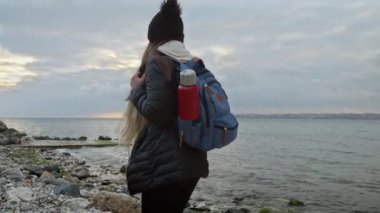 Sırt çantalı ve termoslu genç bir kadın kayalık deniz sahilinde yürüyor, serin esintinin ve dalgaların sesinin tadını çıkarıyor. Macera, keşif ve seyahat kavramı