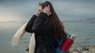 Sırt çantalı gülümseyen bir kadın, soğuk, rüzgarlı bir sahilde yürüyüş yaparken kulaklıkla müzik dinliyor. Maceranın, seyahatin ve müziğin neşesinin özünü mükemmel bir şekilde yakalıyor ve seyahat videoları için ideal hale getiriyor..