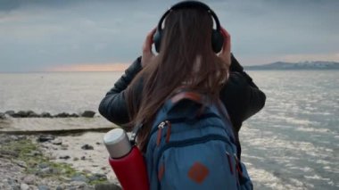 Sırt çantalı gülümseyen kadın deniz sahilinde kulaklıklarını çıkartıyor ve sakin, soğuk okyanus dalgalarına bakıyor. Seyahat kavramı, yolculuk, müzik zevki