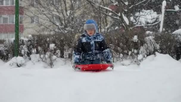 若い男の子が雪に乗ってスローモーションで丘をそります 笑いと喜び冬の風景の美しさ — ストック動画