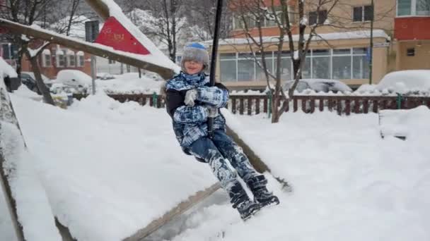 公園で冬にジップラインに乗る陽気な笑顔の少年のスローモーション 冬休みや週末に屋外で遊んだり楽しんだりする子供たち — ストック動画