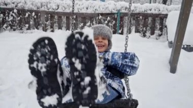 Kar fırtınası ya da kar fırtınası sırasında salıncakta sallanan sevimli gülümseyen çocuk. Kış tatillerinde eğlence ve neşe, dışarıda oynayan çocuklar, kar yağışında faaliyet